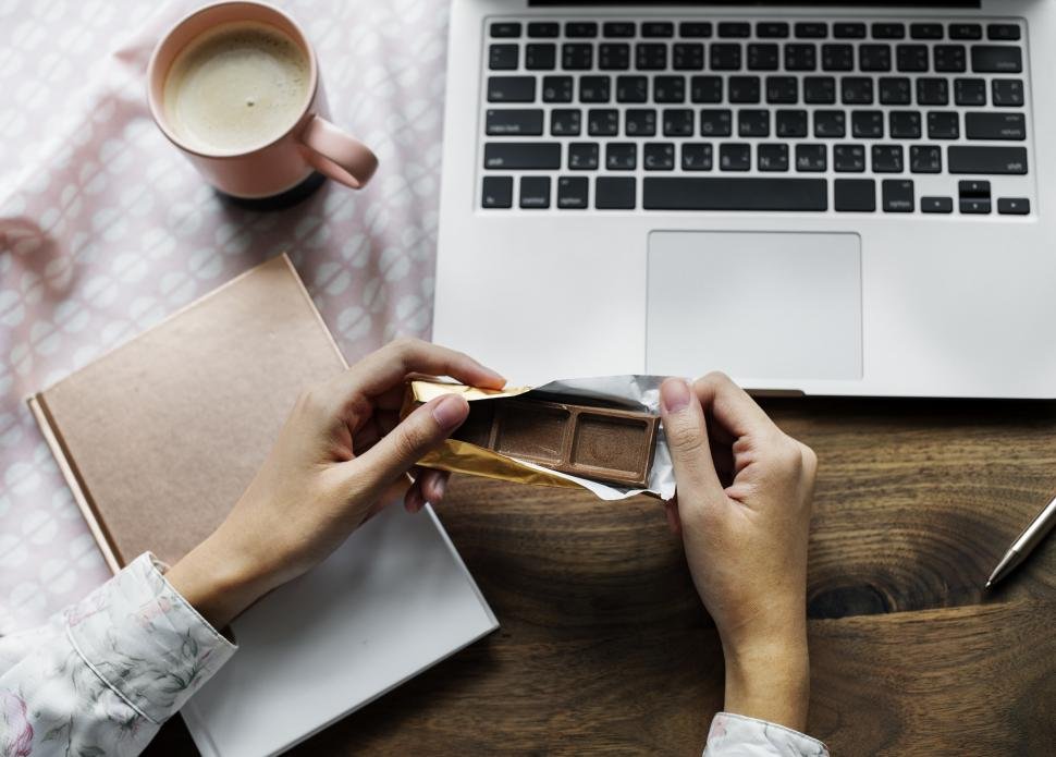mãos sobre a mesa, próximas ao desktop, acompanhada de xícara de café e barra de chocolate.